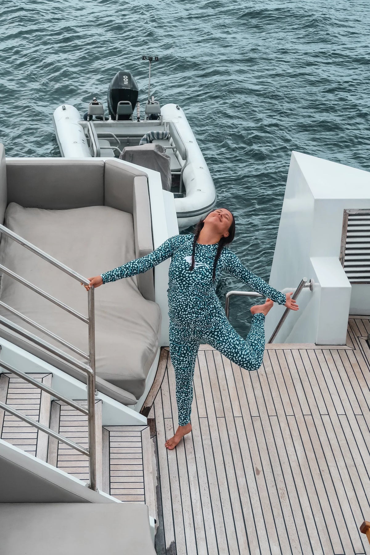 A woman in a whale shark
patterned one-piece swimsuit or bodysuit stretches on the wooden deck of a yacht next to the blue water, with a white inflatable boat in the background.