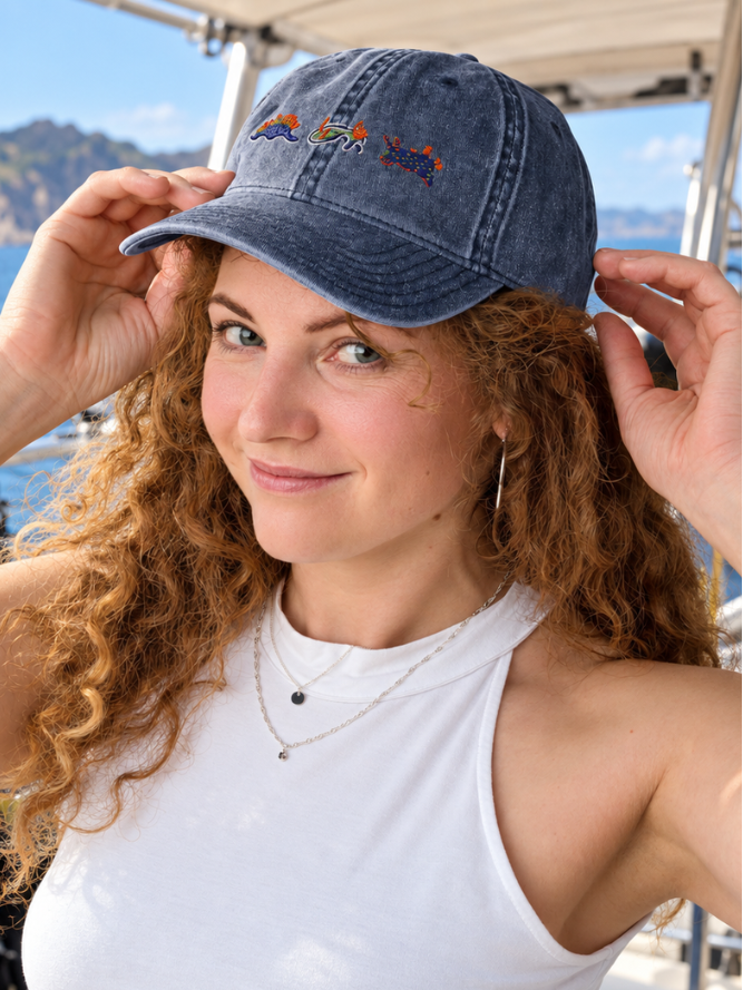 A woman with curly red hair wearing a blue denim baseball cap with a colorful embroidered nudibranch logo, posing on a boat with the sea and mountains in the background.