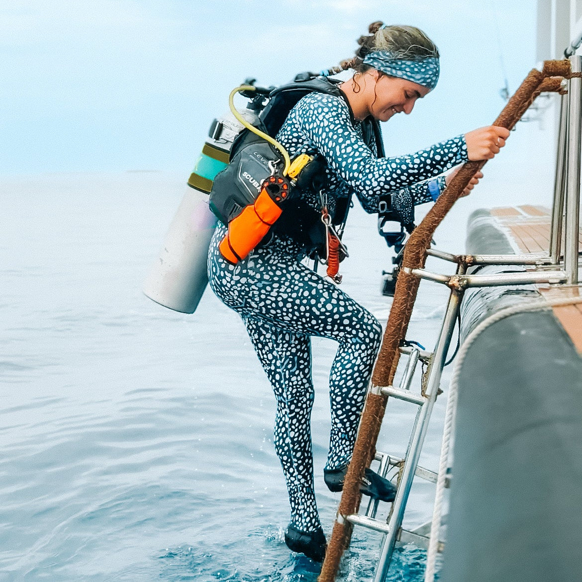 A woman in a blue and white spotted wetsuit climbs a metal ladder off a boat into the ocean, wearing scuba gear with an oxygen tank.